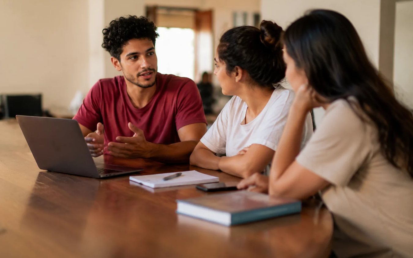 Três participantes conversam ao redor de uma mesa comprida numa sala clara de janelas altas